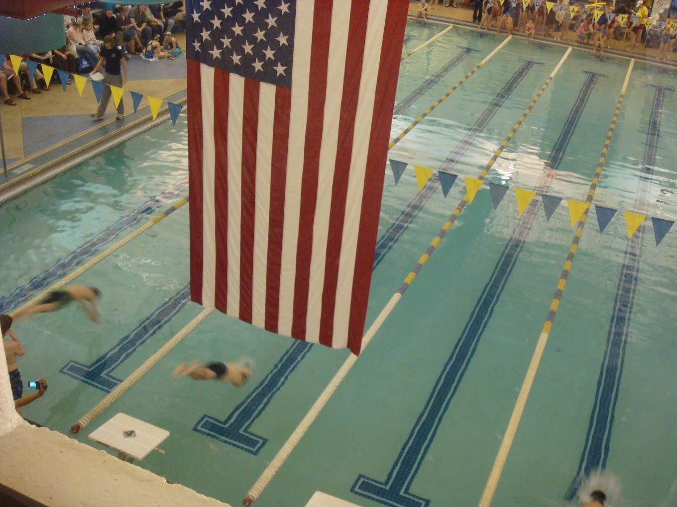 An view of a pool with many lanes for swimming, banners line the pool along with an American flag being flown.