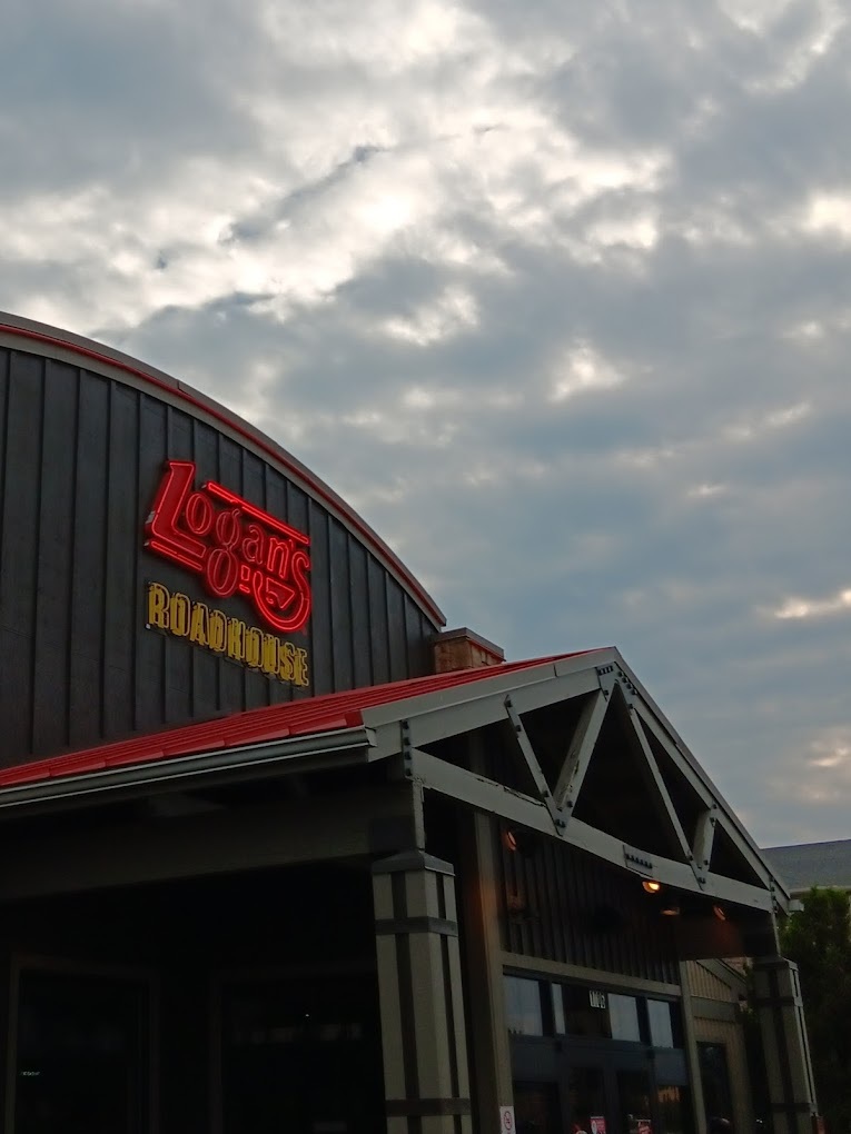 An view of a very large restaurant building with green siding and a large awning above the entrance. A large neon sign in red that read "Logan's Roadhouse"