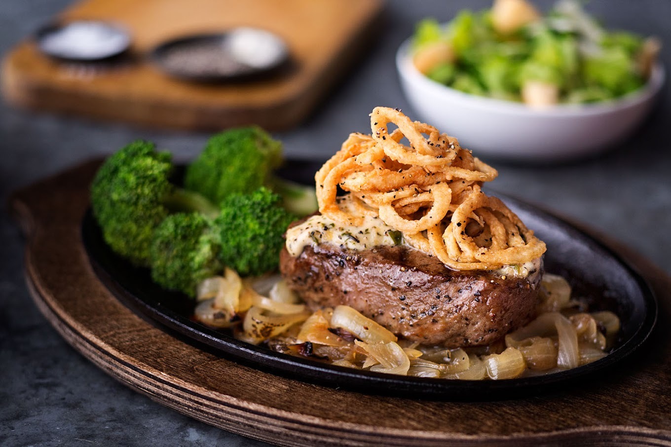 A wooden serving tray with a black plate a top consisting a bed of onion with a steak and topped with onion rings, with a side of lettuce.