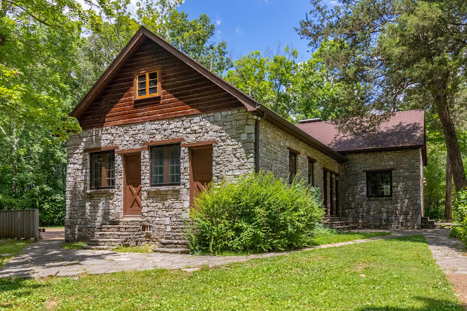 An view of a house surrounded by trees, the house is a cabin style with rocks and dark wood.