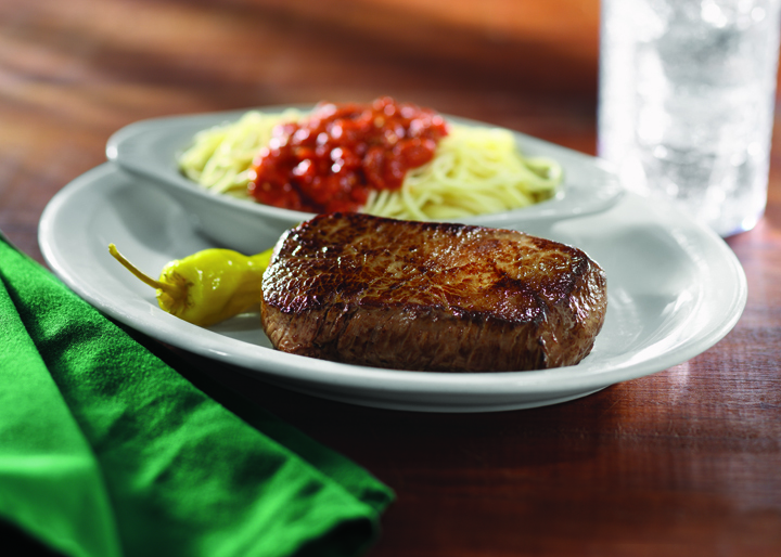 A white plate with a steak, a pepper on the side and a side dish in a white bowl of spaghettis and sauce.