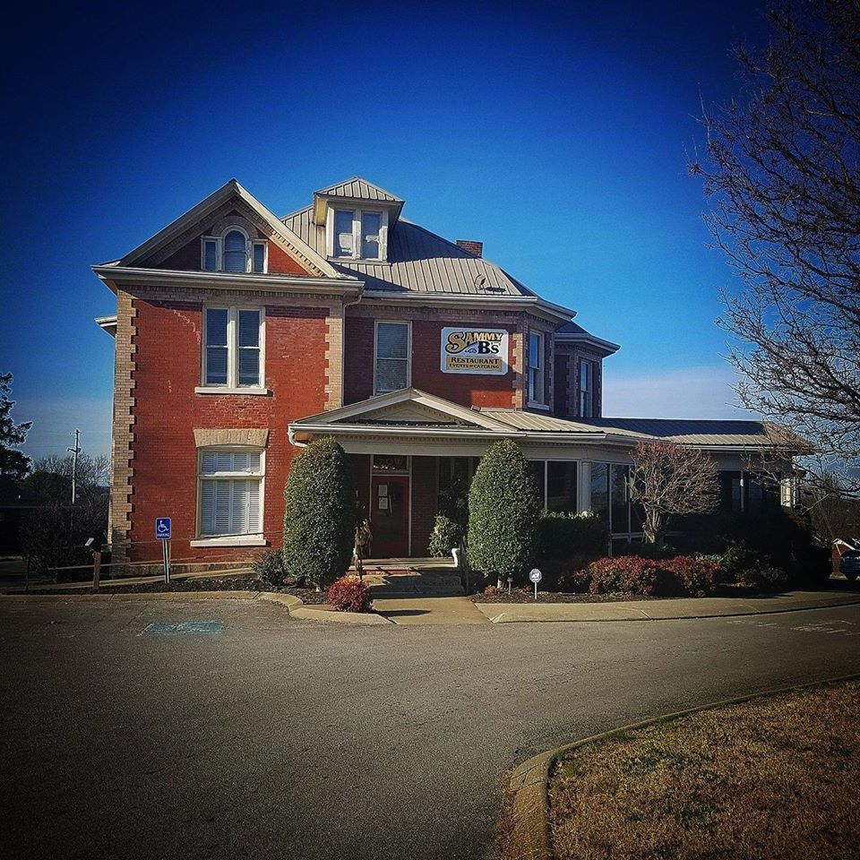 An view of a red building with shrubbery around the house. A yellow sign by the door reads "Sammy B's"
