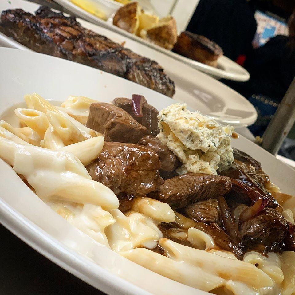 A view of a white plate full of fettucine, topped with sliced steak and topped with some butter with herbs.