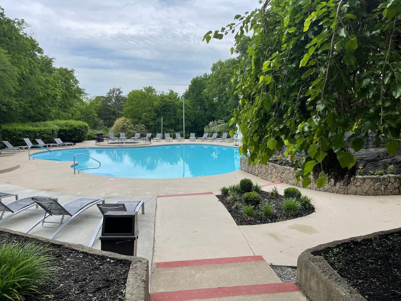 An outside view of a round shaped in ground pool surrounded by bushes and trees, lots of lounge chairs line the side of the pool.