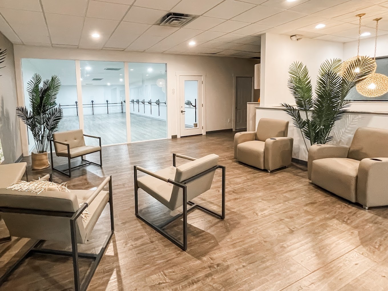 A very bright and modern themed waiting room with a glass window and door leading to a large white dance room with balance bars lining the walls.