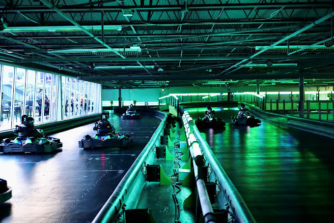 Windows are letting in white light on the left side. Inside, it is dark and green and blue lit. The area is a go karting road, with at least five karts driving.