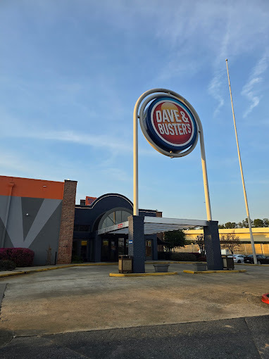 Exterior view of a building with a long sunshade and a large circular entrance sign. It is yellow and the center sign is blue and red. It says "Dave and Busters."