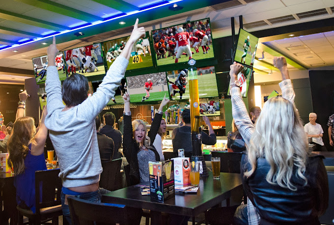 People gathered around dark tables and chairs, as well as a bar table. Their hands are raised in the air and they have excited expressions on their faces, due to winning football game that is shown on the tv screens by the bar table.