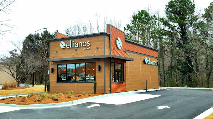Exterior corner view of a an orange and red building with dark accenting, and orange rock landscaping. There is a road with arrows pointing the right direction, and the background has green trees.