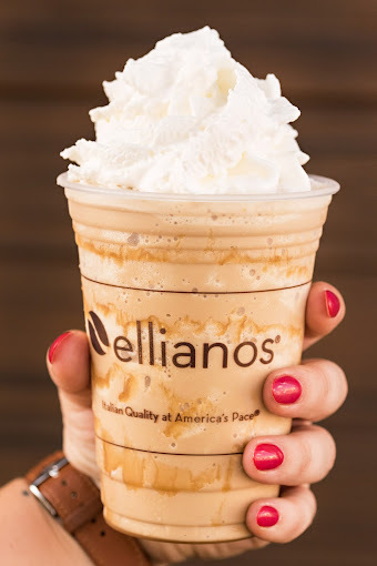 Hand with red nail polish and brown watch holding an iced coffee with whipped cream. The logo on the plastic cup reads "Ellianos."
