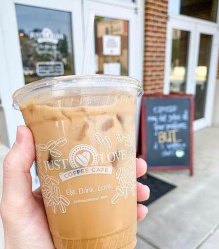Hand holding an iced coffee in a plastic cup, with the background being a brick building with white framed doors. The text on the cup is white and delicate, featuring the name "Just Love" and their slogan.