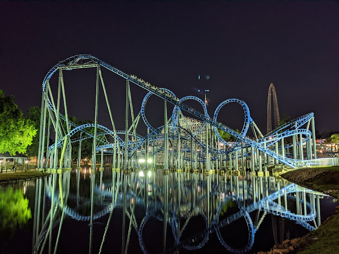 Nighttime view of a blue and green glowing rollercoaster that goes over a lake. The rollercoaster has a reflection in the water.