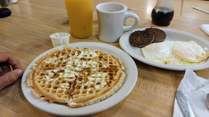 Two dishes of waffles, eggs, and patties on white plates, on a brown wooden table. Also on the table are containers of syrup, a cup of orange juice, and a white coffee mug.