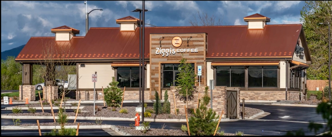 Exterior view of a long red-roofed, beige, and brick based building. Their are drive through lanes with rock landscaping and small green bushes and trees.