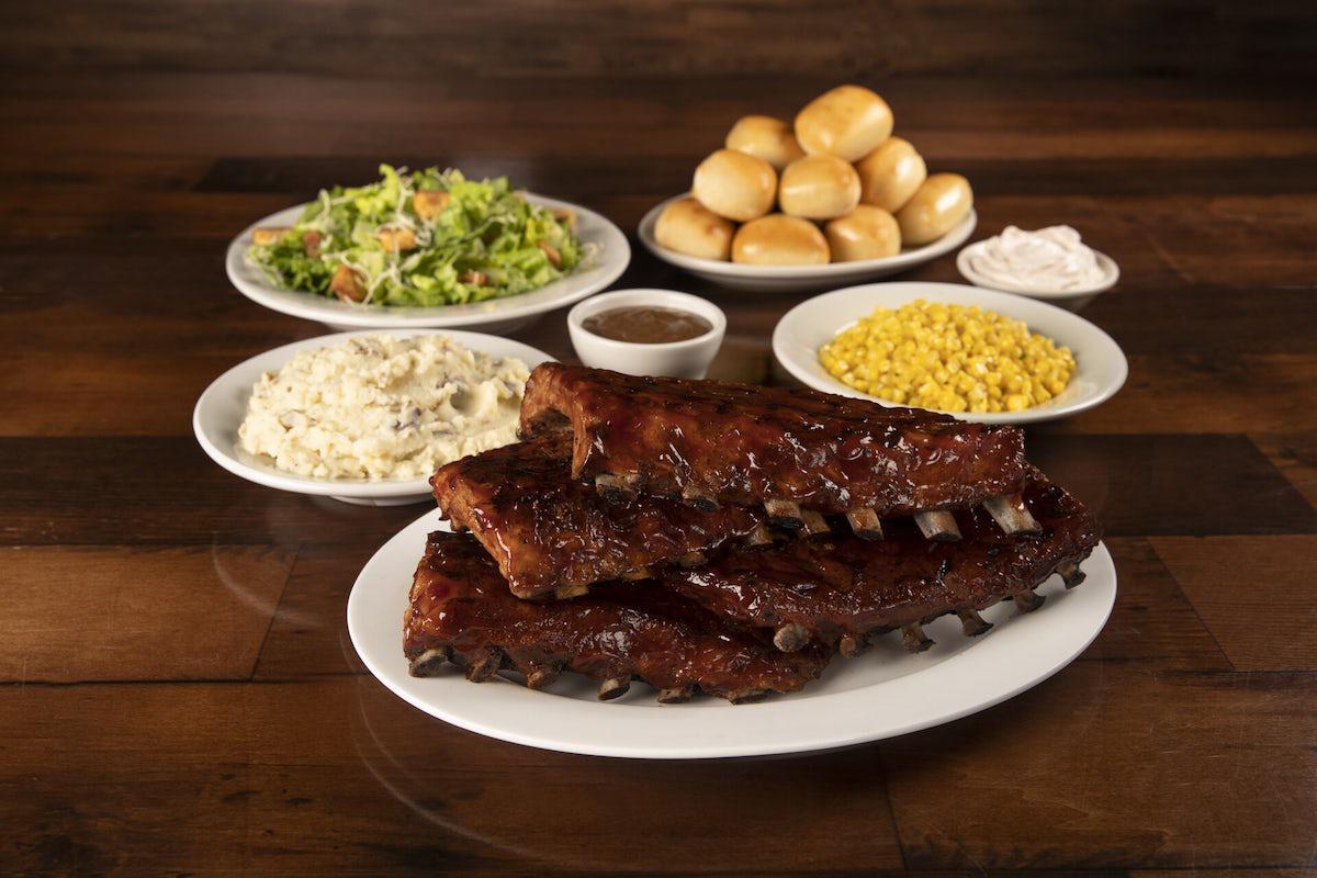 Dinner setting on table with many white plates of food, a rack of ribs, coleslaw, corn, gravy, bread rolls with bread.