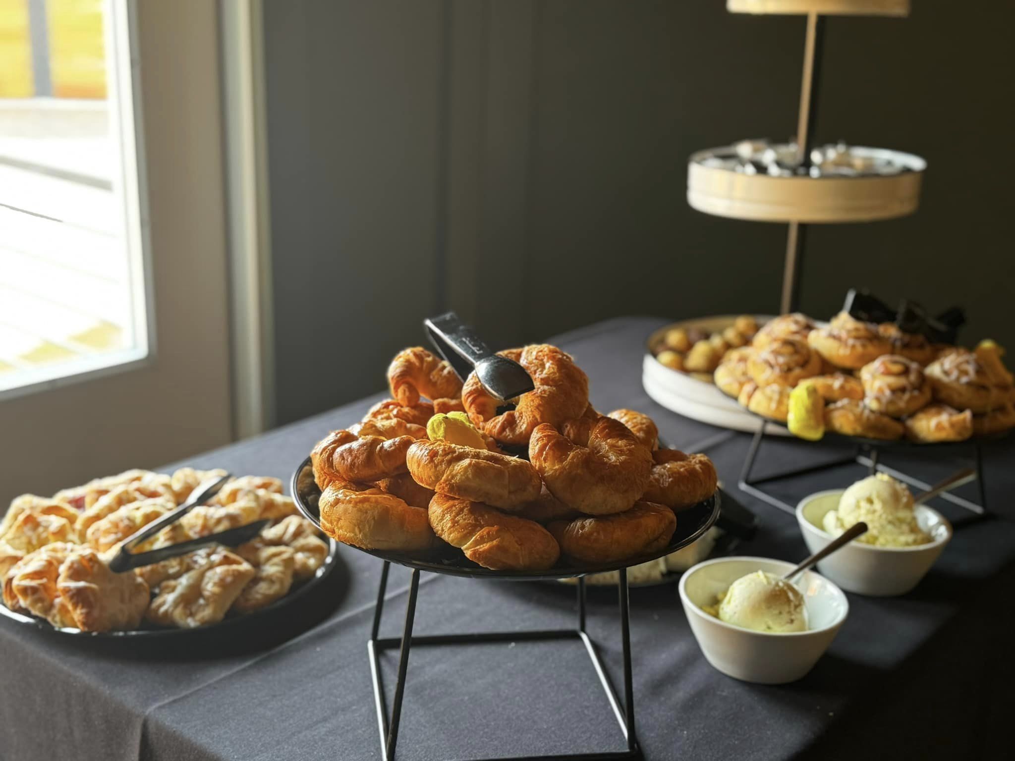 Table with black table cloth covered in various baked goods. The main focal is a risen plate of croissants. The background contains a three-tiered plate of baked goods.