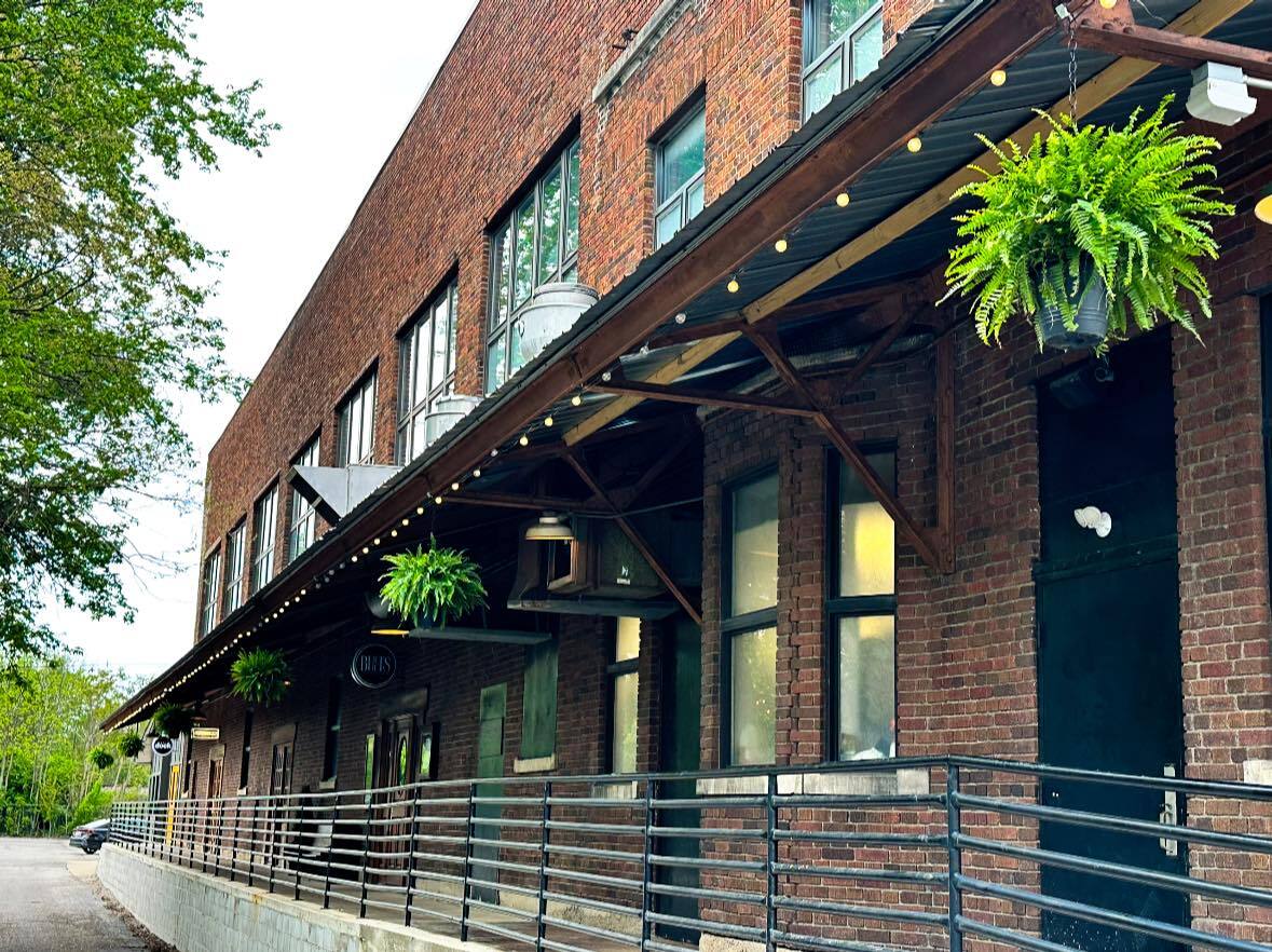 Brick building with a long sunshade stabilized by wooden posts. Along the edge of the building is a risen sidewalk and fence.