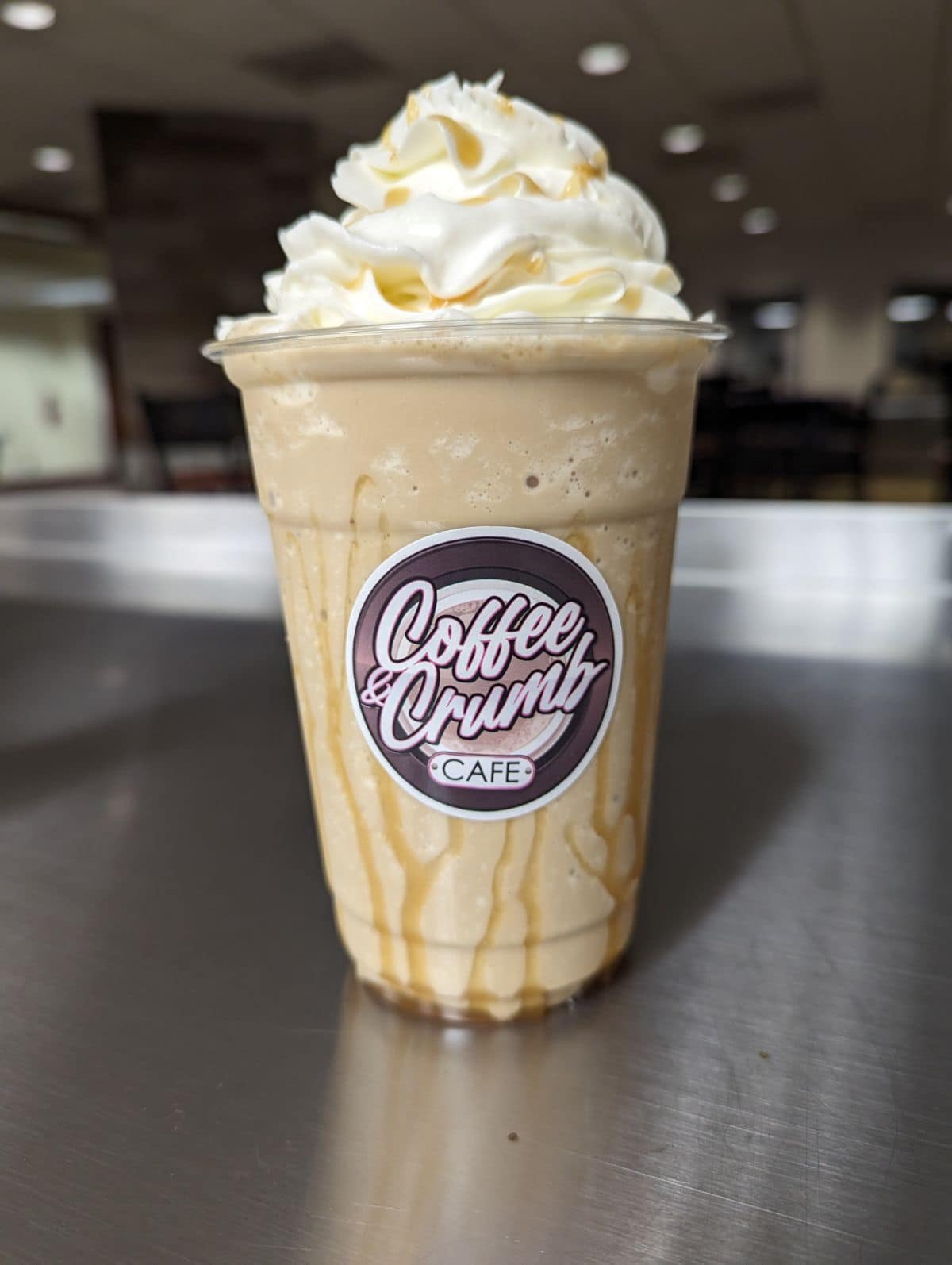 Blended ice coffee with caramel drizzle and whipped cream in a plastic to-go cup. The logo on the front is purple and circular. It says "Coffee and Crumb Cafe."