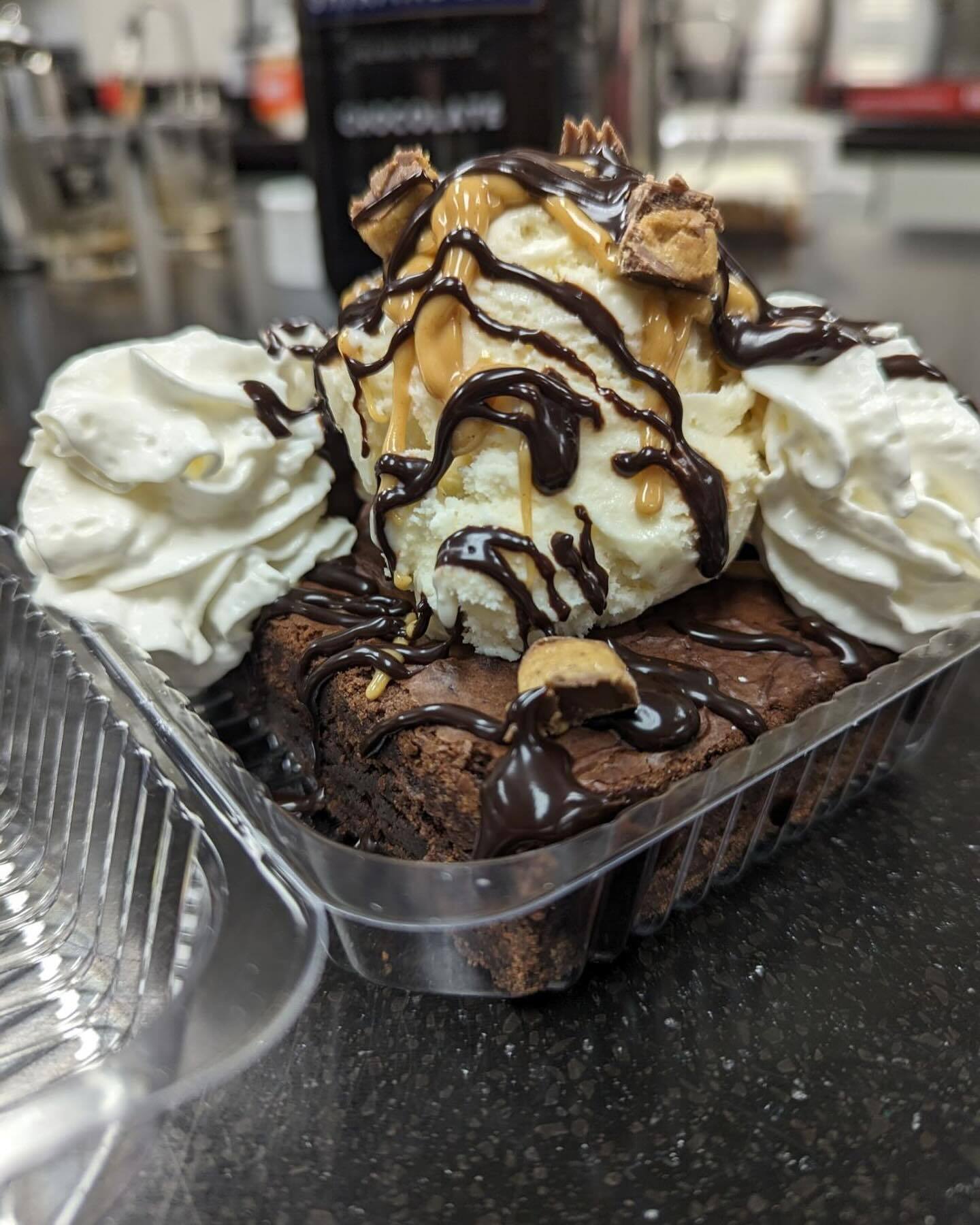 Chocolate brownie cake with ice cream and whipped cream drizzled with caramel and chocolate. This is in a plastic to-go box.
