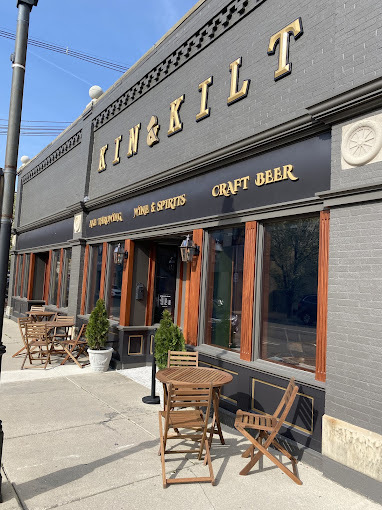 Exterior front view of a building in a downtown setting. There are wooden accents and the signage is gold-toned. There are two round wooden tables accompanied by three chairs each and a plant.