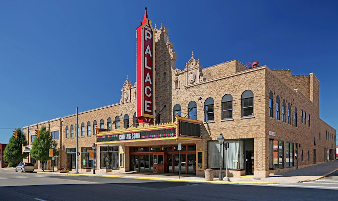 Exterior corner view of a large brick building with two rows of arched windows. The entrance has a large vertical sign that extends tall than the building roof. It's red and it reads "palace."