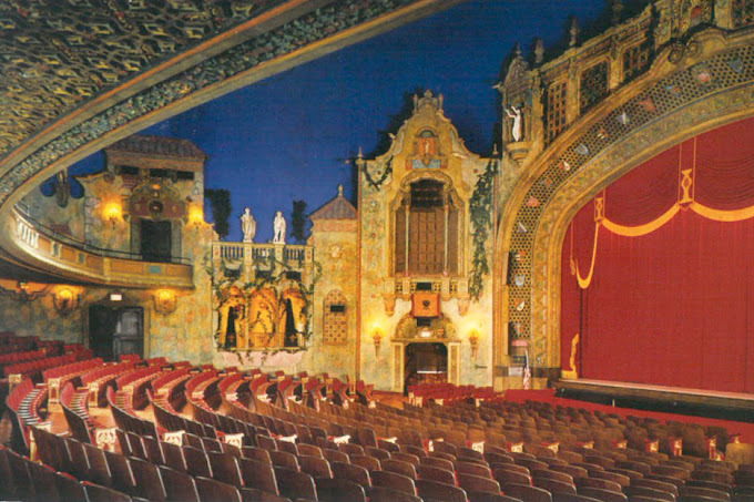 Interior of a theater room. It is ornately decorated and has red seats and curtains. There is a balcony layer above the ground-floor seating.