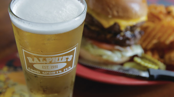 Close up image focused on an alcoholic beverage in a glass cup, with a plate containing a burger and fries is in the background.