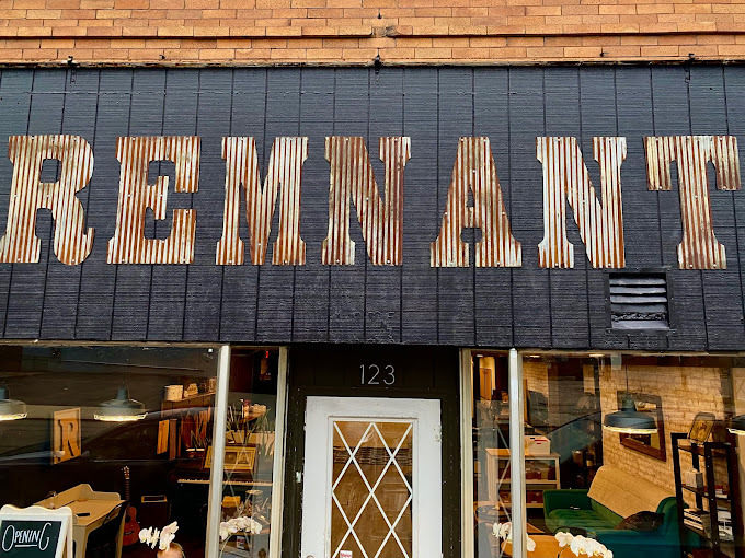 Close view of a building entrance. The sign above the door reads "Remnant" in wooden lettering. The door entrance has white lattice on it, and the windows on either side show the interior, which features a long green couch.