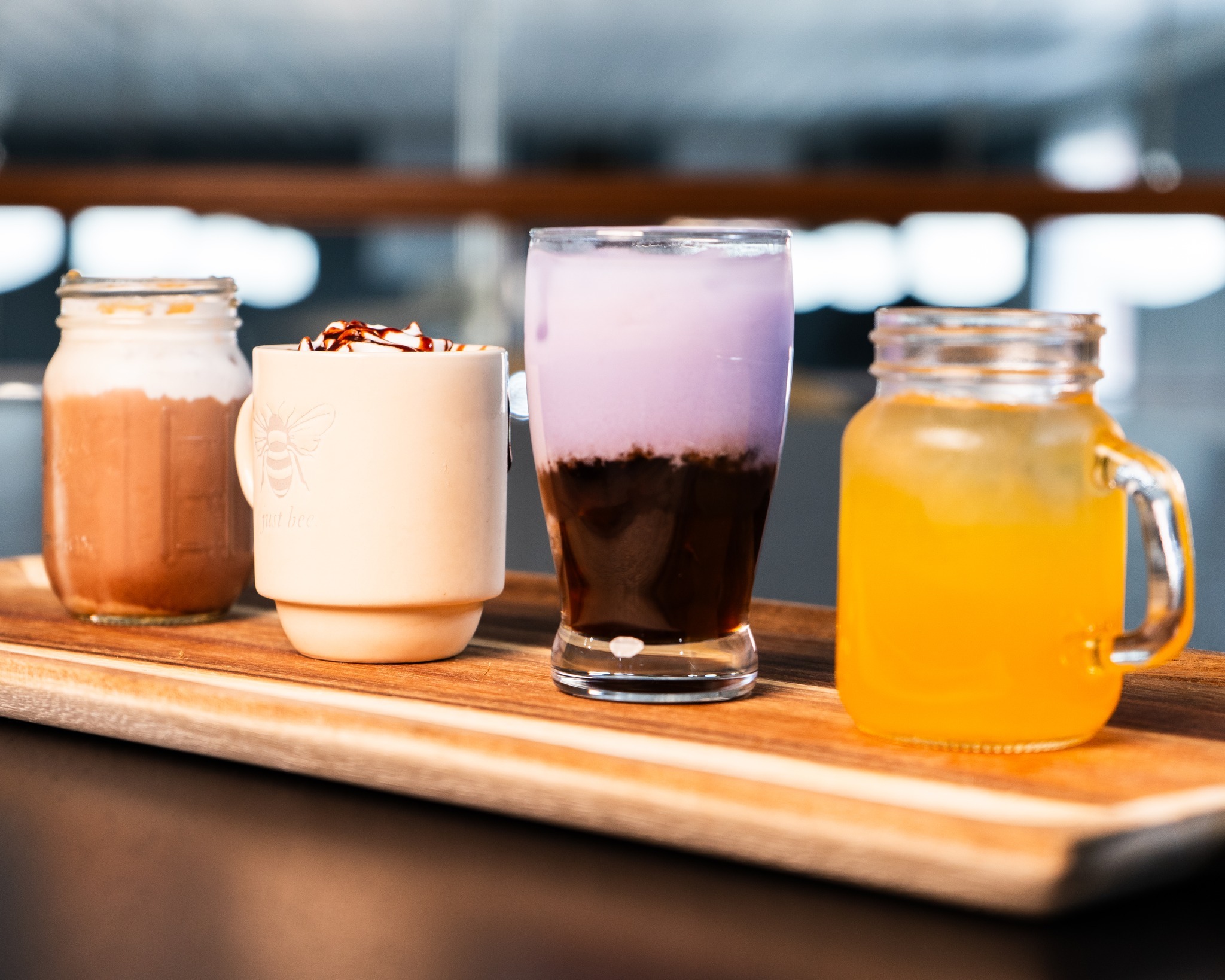 There are four drinks lined up on a wooden board. From left to right: blended iced coffee with caramel and cool whip, hot latte with cool whip and chocolate drizzle, glass cup containing a purple beverage, and a glass cup with a handle containing an orange drink.