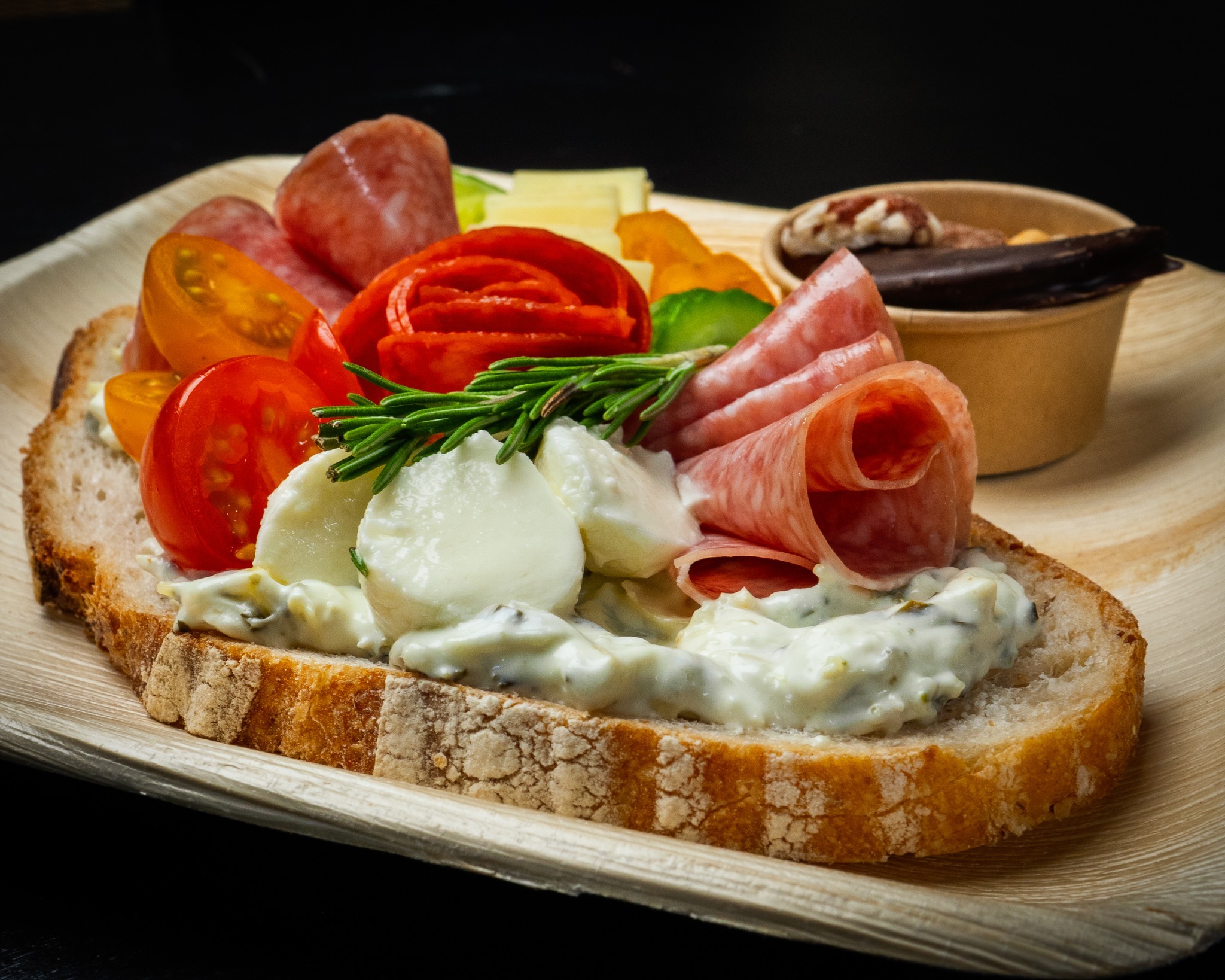 Large piece of bread with deli meat, tomatoes, and cheesy sauce. In the background is a small cup of additional food. This all sets on a rounded wooden plate.