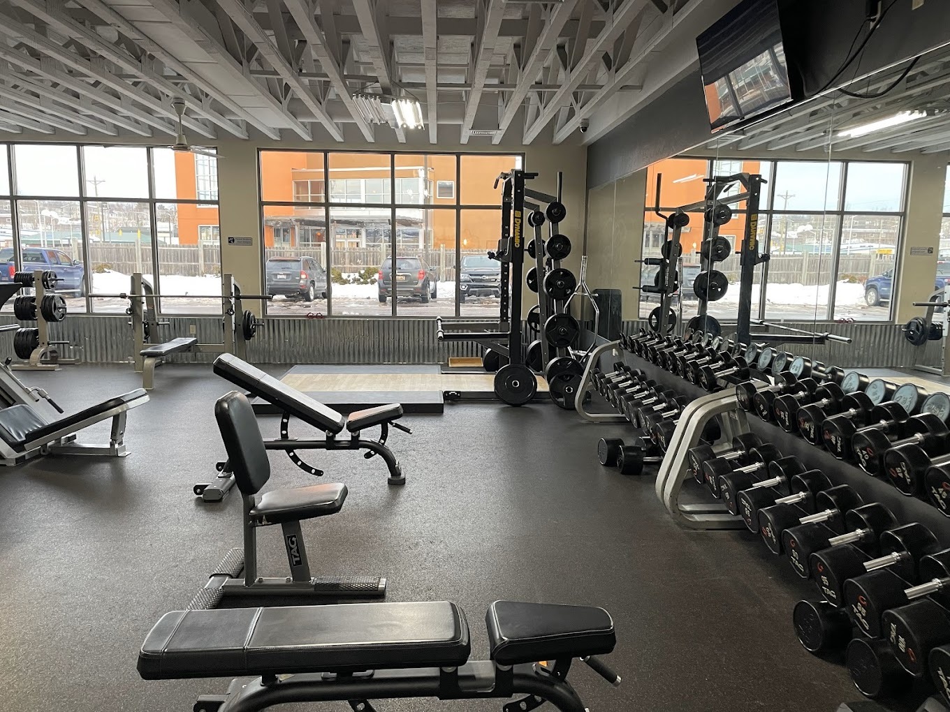 A room with mirrors on the wall, a rack of weights, and lots of benches in a room with large windows.