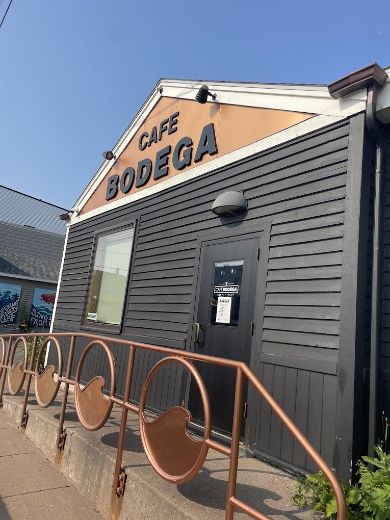 A view of a small building with black siding, tan colored roof with black lettering reading "Café Bodega"