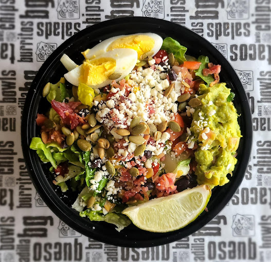 A black bowl consisting of a burrito bowl with rice, lettuce, beans, cheese, guacamole, limes, and egg.