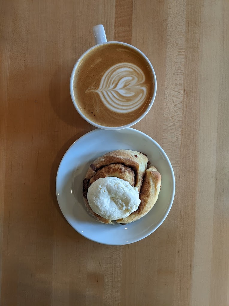 A view of a white coffee cup full of a latte with a spiral latte art along with a white plate with an cinnamon roll.