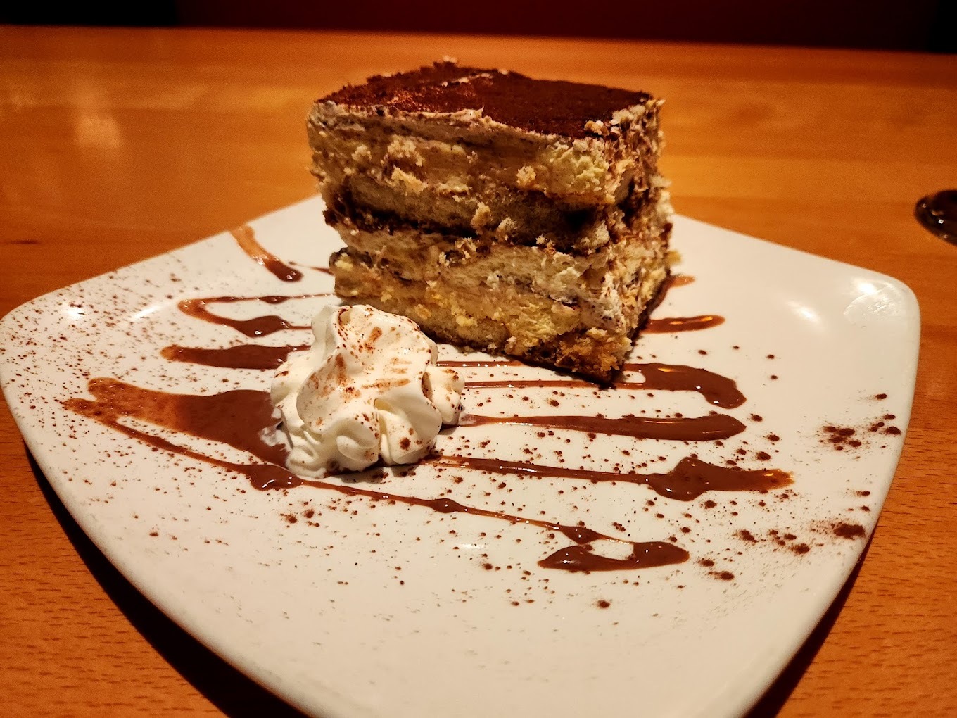A tiramisu cake topped with caramel sauce and cocoa powder, with a side dollop of whipped cream and striped design made of caramel on the plate.