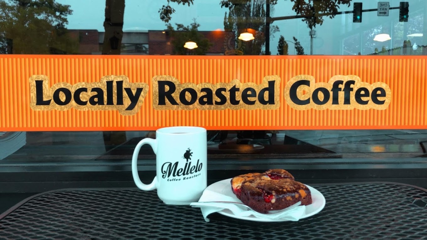 An view from outside on outdoor seating a cup of coffee and pastry on a plate in front of a decal that reads "Locally Roasted Coffee" on an orange decal.