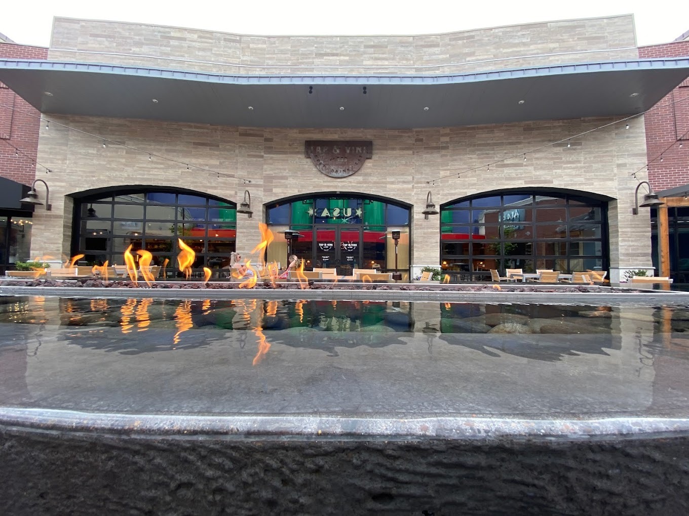 An outside view of a restaurant with outdoor seating, an water feature in the middle of the plaza with a fireplace inside.