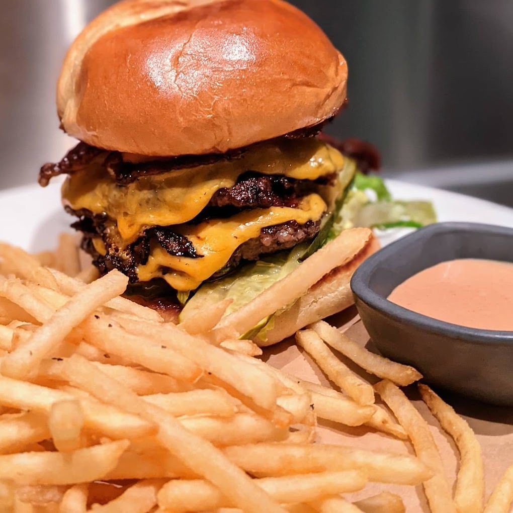 An view of an large stacked burger with a side of classic cut fries and a side of orange sauce in a blue cup.