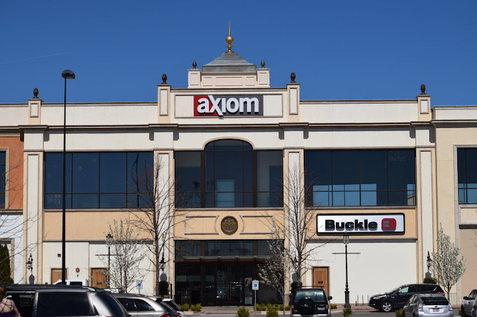 Exterior view of a light colored building with square columns and large windows. There are a few small trees next to the entrance. A sign at the top of the building reads "Axiom." Another sign halfway down the building reads "Buckle."