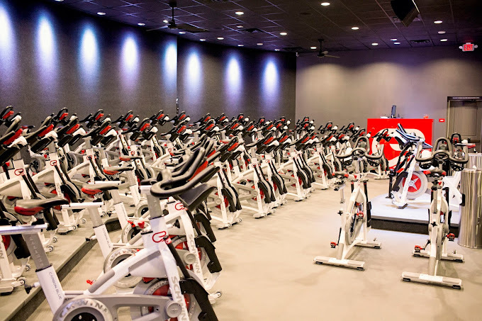 Stationary red, grey, and white bicycles lined up in rows. Towards the front of the room is a stationary bike on a podium.