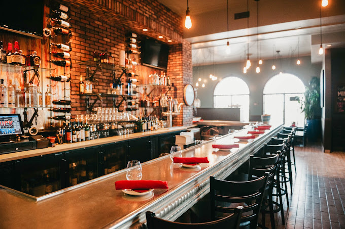 View of a bar table with brick walls, bottles, and two hanging TVs. The far background has arched windows with lots of sunshine.