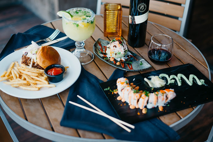 Two sushi plates, a plate of burger and fries, and two drinks on a wooden table.