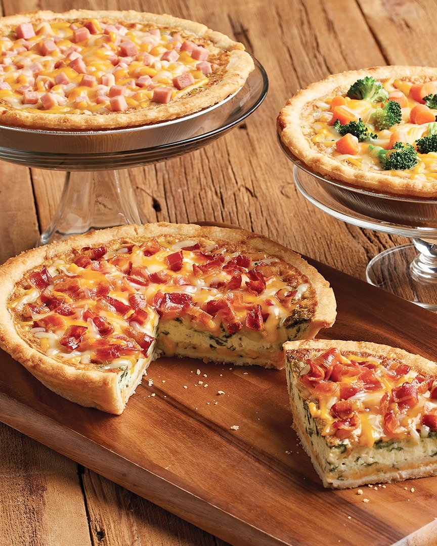 Three different breakfast pies, featuring ham, broccoli, and bacon. Two are set on platter stands, and the other is set on a wooden board, with a slice cut out.