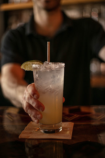 Yellow alcoholic drink with a lemon and straw on a napkin. A man in a black polo holds the side of the drink.