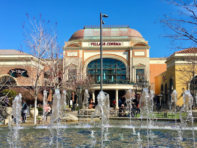 Light colored building with arches, a large window, circular balcony, and columns. The window has lights strung across it, and the balcony holds teal umbrella tables. The top of the roof is turning blue. The front of the entrance features a large water fountain.