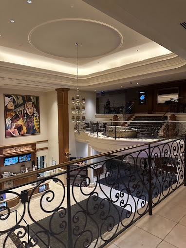 Interior view from a balcony. There is a black swirl fence along the balcony edge. The ceiling is raised and has a small chandelier hanging. The movie theater lobby desk can be seen.