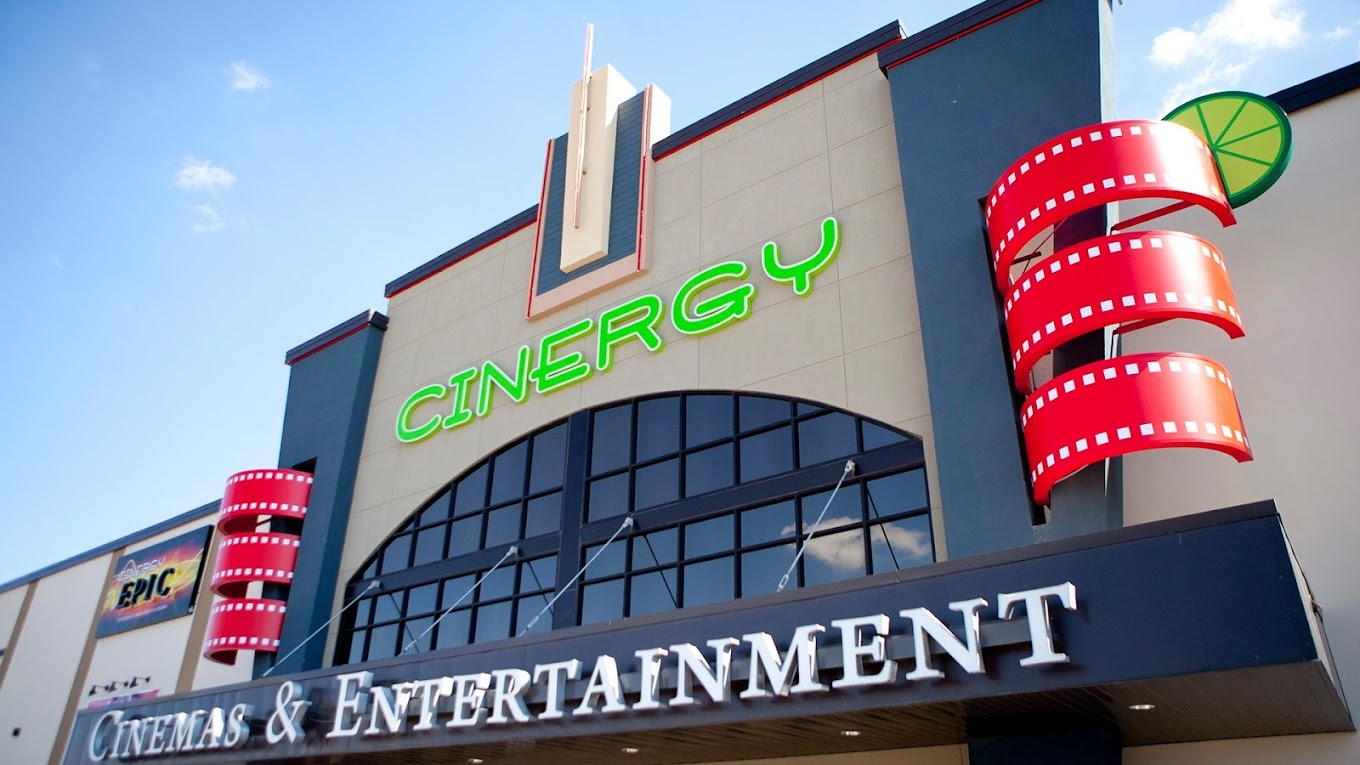 An outside view of a theatre, a large beige building with green lettering on the front of the building above the doors.