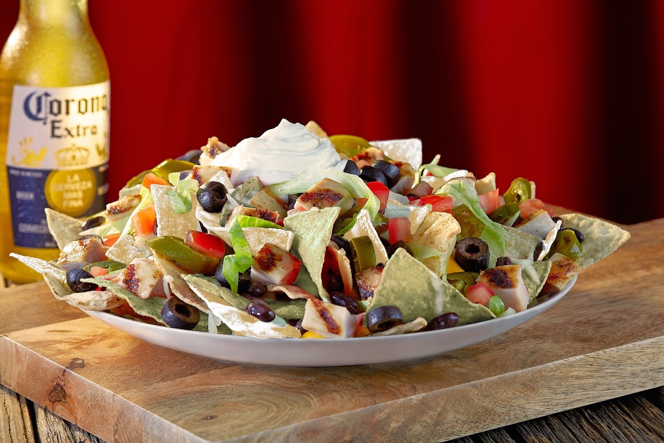 A view of nachos setting on a wooden serving tray with a bottle of beer in the background.