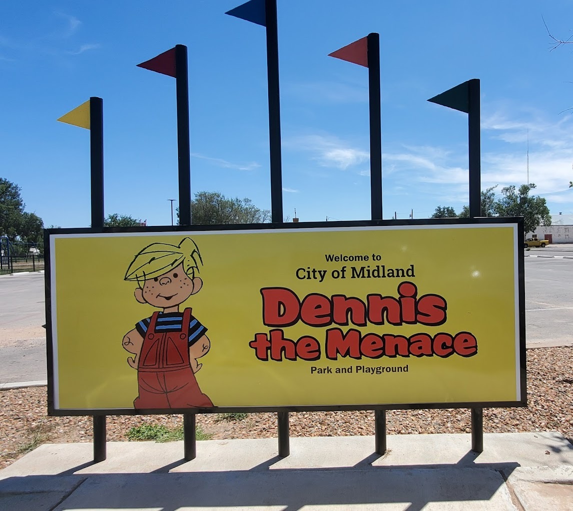 A view of a sign outside of the park area, a yellow sign with colorful flags on the top of the sign. The sign reads "Dennis the Menace"