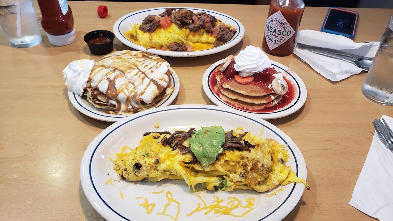 A view of a table full of white plates with different entrees like an omlette, berry pancakes, and smores pancakes.